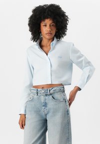 Light blue cropped button-up shirt with a collar, featuring a subtle striped pattern and embroidered logo. Paired with high-waisted light denim jeans.