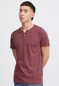 Camiseta de algodón en color marrón con escote henley y tapeta de tres botones. Mangas cortas y un pequeño parche con el logo en la parte inferior izquierda.