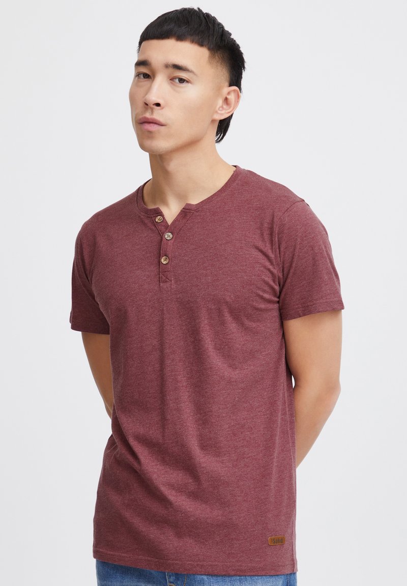 Camiseta de algodón en color marrón con escote henley y tapeta de tres botones. Mangas cortas y un pequeño parche con el logo en la parte inferior izquierda.