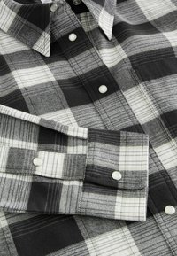 Chemise à carreaux noire, blanche et grise avec des boutons pression et un poignet plié, montrant une partie du col et de la poche poitrine.