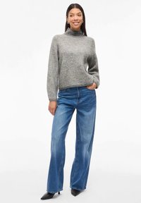 Γυναικα που στέκεται με το ένα χέρι στην τσέπη, φορώντας γκρι ζακέτα με σχέδιο, μπλε φαρδιές παντελόνες και μαύρες μυτερές γόβες.