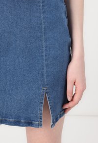 Kék farmer szoknya, slim fit szabással, rövid hasítékkal az alján és kontrasztos sárga varrással az oldalvarrások mentén. Részben látható kéz.