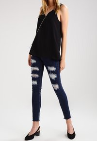 Top negro sin mangas con un diseño fluido, combinado con unos jeans ajustados azul oscuro que presentan varios parches desgastados y un dobladillo ligeramente deshilachado.