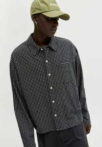 Persona con una camisa de cuadros en blanco y negro, pantalones negros y una gorra de béisbol verde claro, mirando hacia abajo a un lado.