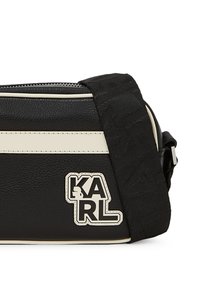 Sac à bandoulière noir en synthétique avec des accents crème, surface texturée, sangle ajustable et patch logo "KARL" à l'avant.