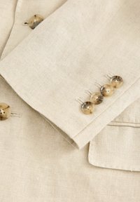 Détail d'une veste en lin beige présentant un poignet replié avec quatre boutons marron clair et des accents cousus. Texture lisse avec un léger éclat.