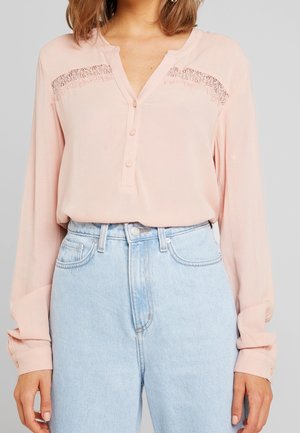Frau trägt eine hellrosa Langarmbluse mit Spitzendetails und hellblaue, hoch taillierte Jeans.