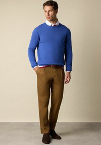 Modrý svetr s kulatým výstřihem, hnědé chinos kalhoty a tmavě hnědé boty. Viditelný bílý límec a červený akcent na košili pod ním. Hladká textura.