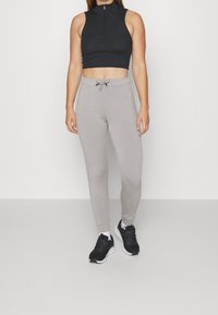 Top corto negro con cremallera y pantalones de chándal gris claro con bolsillos laterales, combinados con zapatillas deportivas negras, de pie sobre un fondo neutro.
