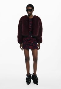 Chaqueta bomber de piel sintética burdeos, con cierre de cremallera, sobre un vestido ajustado con un patrón floral; modelo luciendo gafas de sol negras de gran tamaño y botas escultóricas.
