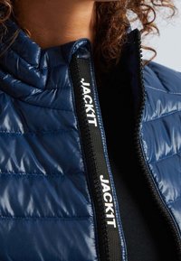 Laivastonsininen tikattu untuvatakki vetoketjulla. Mustassa vetoketjunnauhassa lukee "JACKIT", mikä korostaa takin pintaa ja muotoilua.