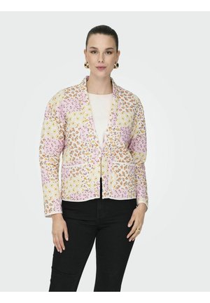 Femme portant une veste patchwork florale pastel par-dessus un haut blanc, associée à un pantalon noir, debout devant un fond clair uni.