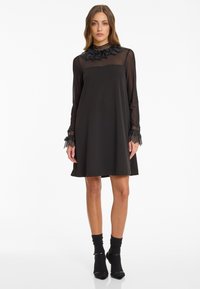 Robe noire avec de longues manches transparentes, un col en dentelle agrémenté d'accents floraux et des poignets texturés. Forme évasée, longueur genou, tissu lisse.