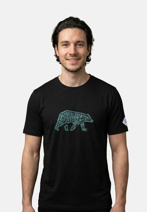 Lachende man met donker haar, gekleed in een zwart t-shirt met een gestileerde blauwe berenafbeelding, loopt naar links op een eenvoudige lichte achtergrond.