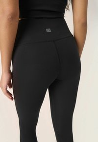 Črne legice z visokim pasom, izdelane iz raztegljivega materiala, z diskretnim logotipom blizu pasu in eleganto gladko teksturo.