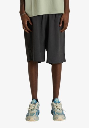 Persona che indossa pantaloncini larghi grigio scuro fino al ginocchio, t-shirt verde chiaro, scarpe da ginnastica blu e beige, in piedi davanti a uno sfondo bianco semplice.