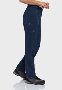 Marine Outdoor-Hosen aus elastischem Material, mit Reißverschlusstaschen und dezentem Logodetail, kombiniert mit dunklen Wanderschuhen.