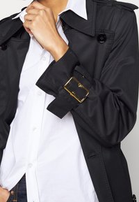 Czarny płaszcz trench z zapięciem na pasek i złotym klamrą, narzucony na białą koszulę. Posiada błyszczące guziki oraz gładką, elegancką fakturę.