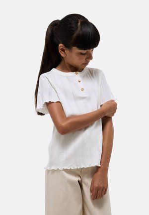 Haut blanc à manches courtes avec col rond, présentant trois boutons et une texture côtelée. Le bord festonné à l'ourlet ajoute du détail.