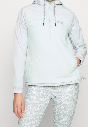 Femme portant une veste à capuche Columbia bleu clair et un pantalon de sport à motifs, les mains dans les poches, debout devant un fond uni.