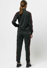 Schwarzes Trainingsanzug mit roten Seitenstreifen, aus glattem Stoff gefertigt. Besitzt einen hohen Kragen und elastische Bündchen an Jacke und Hose.