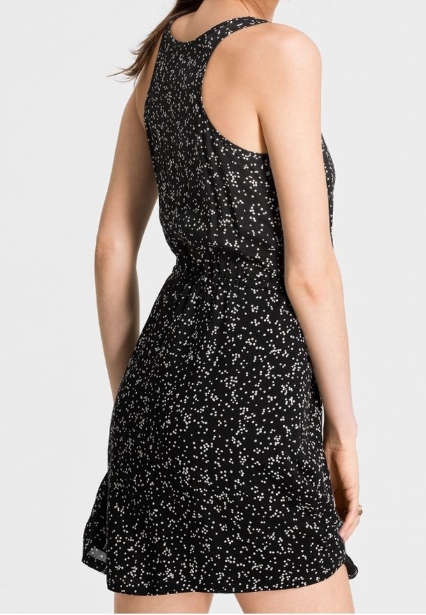 Robe noire sans manches avec un motif moucheté blanc, taille ajustée et style dos nageur, portée par une personne debout vue de dos.