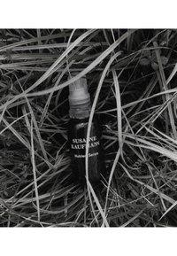 Czarna szklana butelka z pompką, oznaczona napisem "Susanne Kaufmann Nutrient Serum", otoczona zieloną trawą i teksturowanym podłożem.