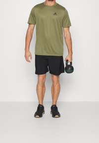 Camiseta deportiva verde oliva, pantalones cortos negros, sosteniendo una kettlebell negra con detalles azules, combinada con zapatillas deportivas negras y suelas de goma.