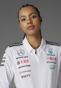 Vit polo t-shirt med svarta ränder, prydd med flera sponsorns logotyper i olika färger på framsidan. Silverfärgad kedja som accent.