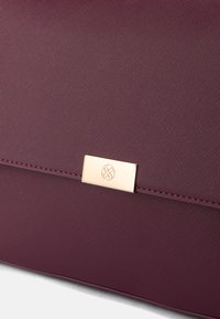 Bordová textúrovaná kabelka s kovovým označením v zlatej farbe a elegantným dizajnom, ktorá sa vyznačuje hladkým povrchom a štruktúrovaným tvarom.