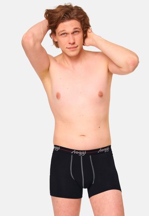 Uomo senza maglietta con pelle chiara e capelli castani che indossa boxer neri Sloggi, posando con le mani dietro la testa davanti a uno sfondo semplice.