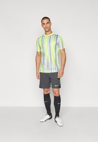Nike Sportbekleidung mit einem neon-gelben und grauen gestreiften kurzärmeligen Shirt, kombiniert mit grauen Shorts, schwarzen Socken und weißen Schraubstollen.