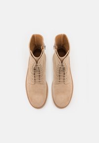 Un paio di stivaletti beige in suede con lacci e cerniere laterali, mostrati dall'alto su uno sfondo bianco.