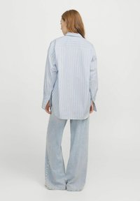 Hemd s dlouhým rukávem v světle modré a bílé pruhované barvě, s špičatým límcem, volným střihem a zaobleným spodním lemem. V kombinaci s širokými džíny z světlého denimu.