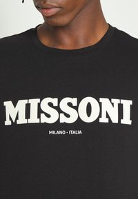 Svart t-shirt med fet vit text som läser "MISSONI" med "MILANO - ITALIA" under i mindre stil. Tillverkad av bomull, rund halsringning.