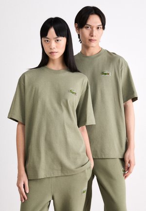 Camisetas de algodón verde olivo con mangas cortas, que presentan un logo de reptil bordado en el lado izquierdo del pecho. Corte casual y textura suave.