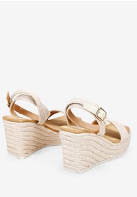 Sandales compensées avec un dessus en textile beige tissé, une bride de cheville réglable et une plateforme en bois naturel. Présente un talon en corde tressée.