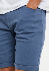 Blå denim shorts med oprevne kanter, med et fladt frontdesign, subtil struktur og sidelommer. Metalaccent nær lommen.