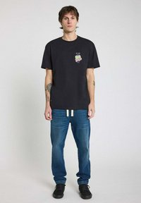 T-shirt en coton noir avec un graphisme coloré "Bad Boy" sur le côté gauche de la poitrine, porté avec un jean en denim bleu et des baskets noires.