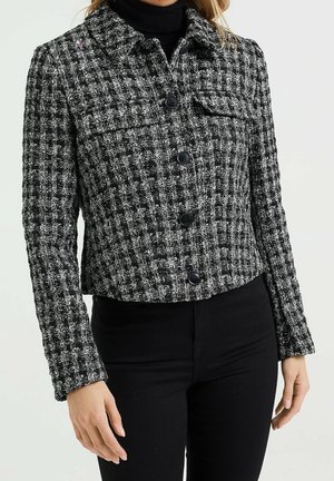 Zwart-witte getextureerde tweed jas met knopen en klepzakken op de borst, gedragen over een zwarte coltrui en zwarte broek.