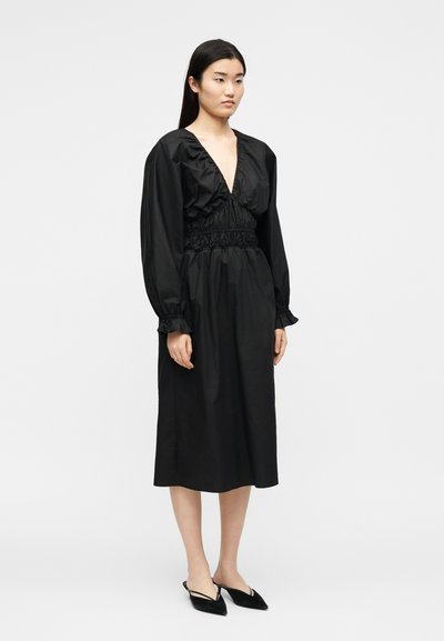 Νεαρή γυναίκα που στέκεται φορώντας μαύρο μίντι φόρεμα με μακριά μανίκια, βαθύ V-λαιμόκοψη, σφιχτή μέση και μαύρα μυτερά παπούτσια με λουράκι πίσω.