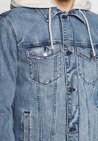 Veste en denim bleu clair avec capuche grise. Comprend deux poches poitrine, des fermetures à bouton et une texture légèrement déchirée. Doublure douce visible.
