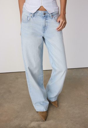 Osoba oblečená v svetlomodrých širokých džínsoch a hnedých semišových topánkach so špicatou špičkou, stojaca na betónovej podlahe s bielym pozadím steny.