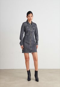 Mørk denim skjortekjole med lange ermer, frontglidelås og to brystlommer. Stylet med svarte ankel støvler. Jevn, tettsittende design.