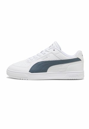 Biały sneaker z niską cholewką, ciemnoniebieskim paskiem z logo Puma, sznurowany przód, perforowany nosek i płaska biała gumowa podeszwa na jednolitym tle.