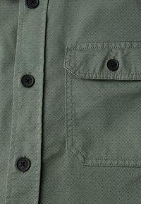 Nærbillede af grøn skjorte foran med sorte knapper og en klaplomme sikret med en knap på højre side.