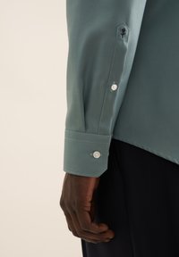 Chemise bleu clair à manches longues, avec des poignets boutonnés, un accent brodé et une texture lisse avec des coutures visibles.