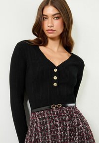 Černý pletený top s V-výstřihem a třemi zlatými knoflíkovými detaily. Modelka ho kombinuje se strukturovanou burgundskou sukní a černým páskem.