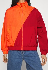 Person som bär en ljus orange och röd färgblockerad zip-jacka med elastiska ärmar och midja, kombinerad med blå jeans.