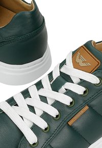 Donkergroene leren sneakers met witte veters, witte zolen, een lichtbruine logo patch op de tong en een "wojas" label aan de zijkant.